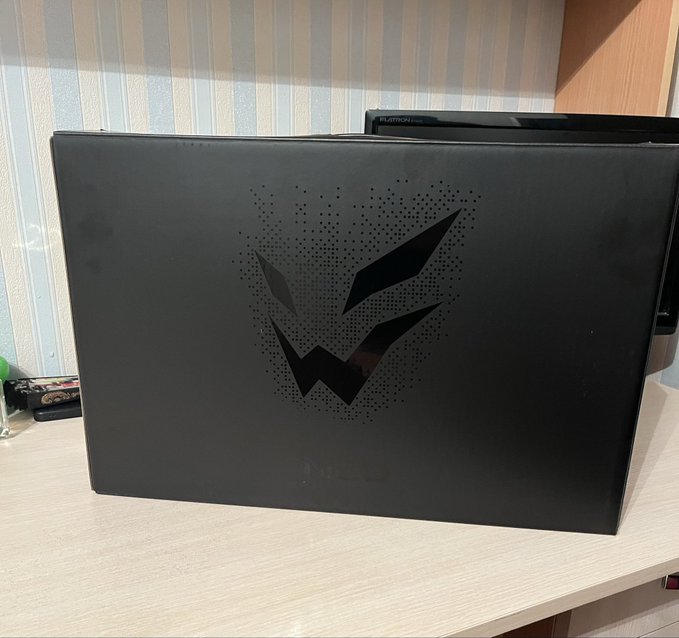 A black laptop box sits on a wooden desk, featuring a stylized, geometric mask logo with dotted shading and the faint 'neo' branding on its lower edge.