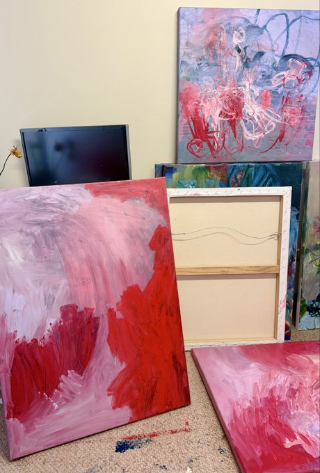 Several abstract paintings with red, pink, and blue brushstrokes leaning against a wall in a room, with one canvas turned around to reveal its wooden stretcher bars.