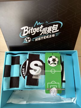 The image features an open box containing a soccer ball and two scarves with a soccer theme. The soccer ball is placed inside the box, while the two scarves are spread out around the ball. The box also contains a small tag attached to one of the scarves. The overall scene suggests that someone has sent these items as a gift or surprise for an avid soccer fan.