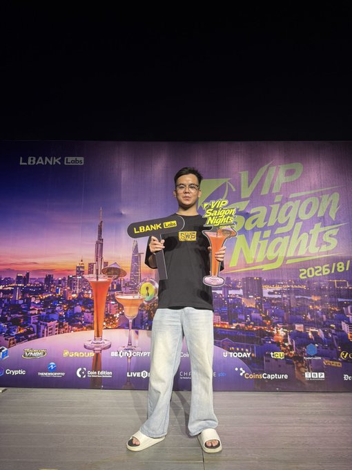 A young man in a black t-shirt, jeans, and slide sandals holding a "LBANK Labs SWE" branded sign and a trophy, posing on stage at the "VIP Saigon Nights" 2026/8/1 event backdrop featuring various crypto sponsor logos.