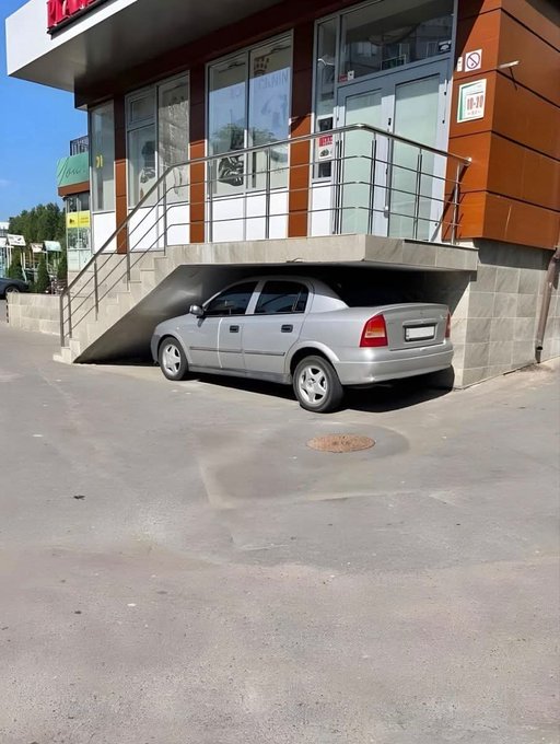 A silver sedan is parked on a paved area beneath a raised staircase leading to a storefront with glass doors.