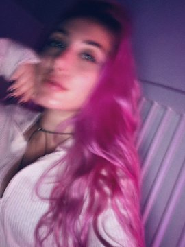 A blurry, purple-tinted selfie of a young woman with vibrant pink hair, wearing a white top.