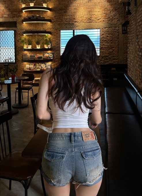 A photo of a woman with long dark hair viewed from behind, wearing a white crop top and denim shorts, standing in what appears to be a coffee shop or restaurant with brick walls and plants.