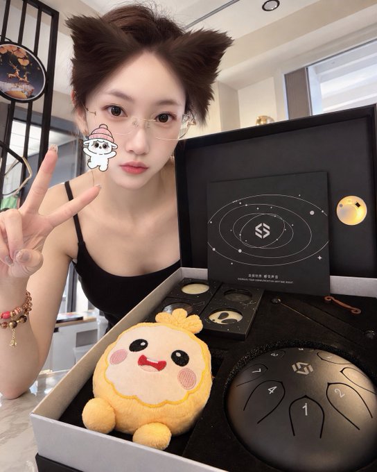 A young woman with short hair wearing glasses making a peace sign, with a small cartoon sticker on her cheek, posing next to a gift box containing a plush toy and a steel tongue drum with a 'SOUND HEAL' booklet.