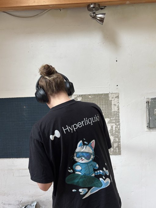 A person with a top knot bun, seen from behind wearing black headphones, sports a black t-shirt featuring a surfing cat graphic and the text 'Hyperliquid'.