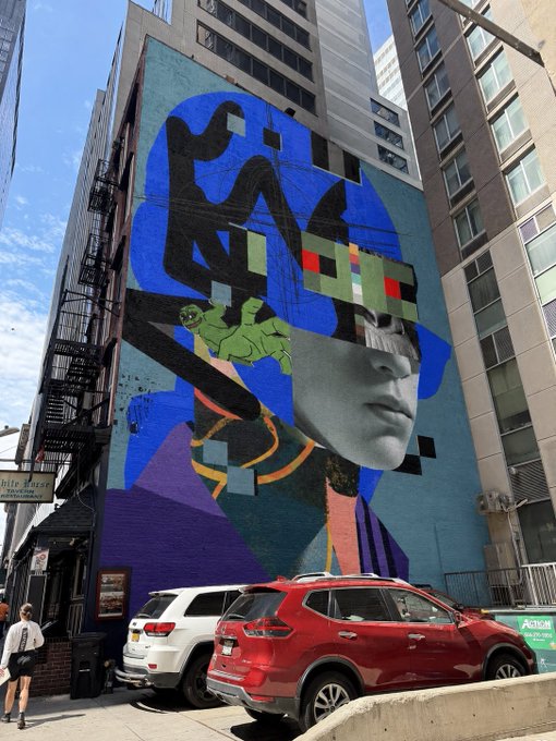A vibrant urban mural painted on the side of a building, depicting a stylized face with a green frog and abstract black shapes against a blue background, with a red SUV parked below.