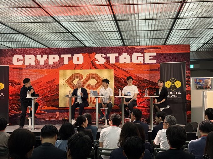 A panel of five speakers sits on stage at the "CRYPTO STAGE" event, with one speaker addressing the audience via microphone while attendees watch from below. Banners for "A" and "IADA NEWS" flank the stage, and a Japanese yen stablecoin logo is displayed behind the panelists, fitting the talk about JPYC's permissionless initiatives.
