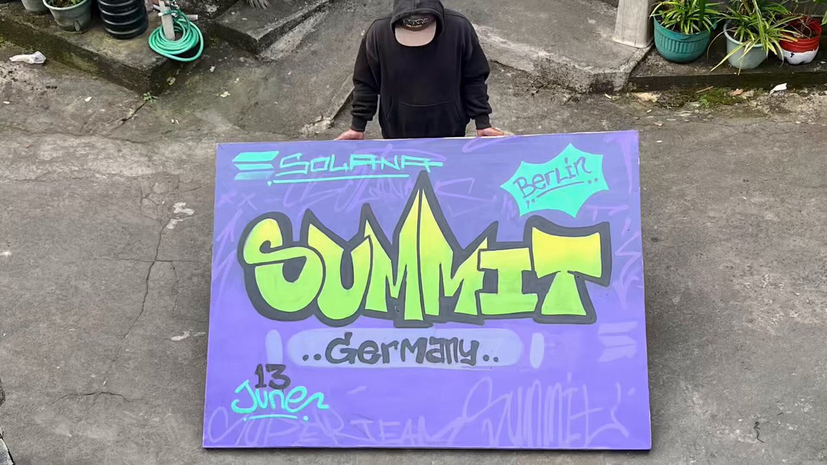 The image features a man holding up a large purple sign with graffiti on it, which says "SOLANIA SUMMIT DIGITAL FESTIVAL". The man appears to be standing outside of a building or on a street, possibly in Berlin Germany as suggested by the date and location information provided. The graffiti on the sign adds an artistic touch to the scene, making it more visually appealing and engaging for viewers.