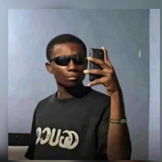 A young man wearing dark sunglasses and a black graphic t-shirt takes a mirror selfie with a smartphone against a gray wall.