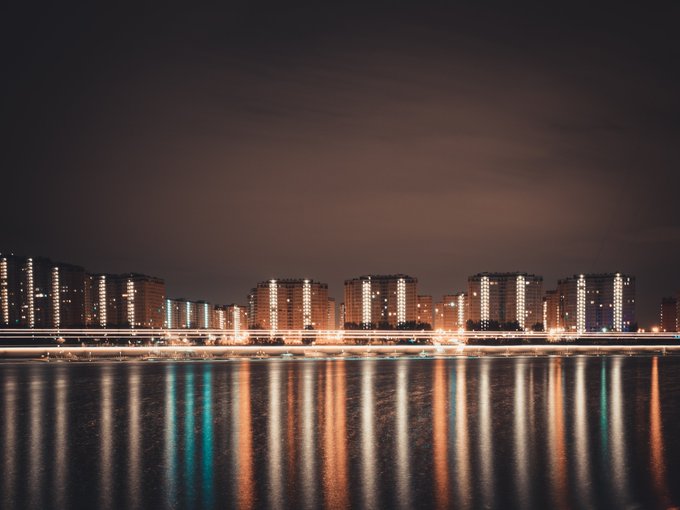 The image shows a nighttime cityscape with tall buildings reflected in the water below, creating a beautiful scene of urban architecture illuminated by artificial light. The long exposure technique used in this photo enhances the brightness and depth of field, making the city lights stand out against the dark sky. The image is captioned "Collection" to indicate that it is part of a collection or series of images related to nighttime city photography.
