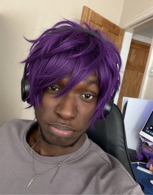 A selfie of a person wearing a purple anime-style wig, large black headphones, and a gray sweatshirt with a silver chain necklace, sitting in a gaming chair.