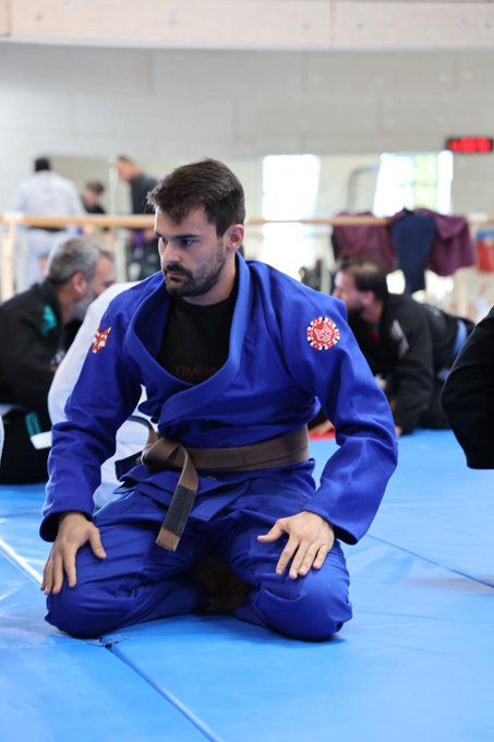 A bearded male athlete in a blue judo gi with a brown belt, kneeling on blue mats during a martial arts competition.
