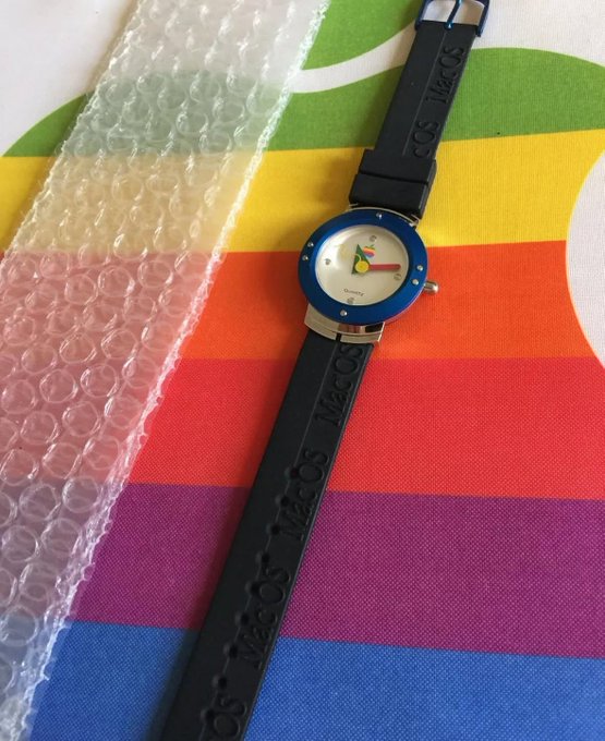 A blue Mac OS watch with a black 'Mac OS' branded silicone strap, resting on a colorful rainbow-striped surface next to bubble wrap packaging.