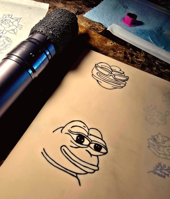 A close-up photo of tattoo sketches on a wooden desk, featuring line-art drawings of cartoon-style creatures with large eyes, alongside a blue tattoo machine with a foam grip and a small container of pink pigment.