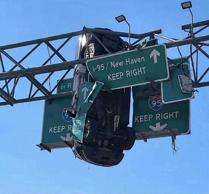 A black Tesla Roadster is stuck upside-down on overhead highway signs for I-95/New Haven, with "KEEP RIGHT" signs visible around the dangling vehicle. The bizarre sight perfectly captures the post's implication of a Tesla stuck hanging from signage, mirroring the feeling of an inescapable financial obligation.