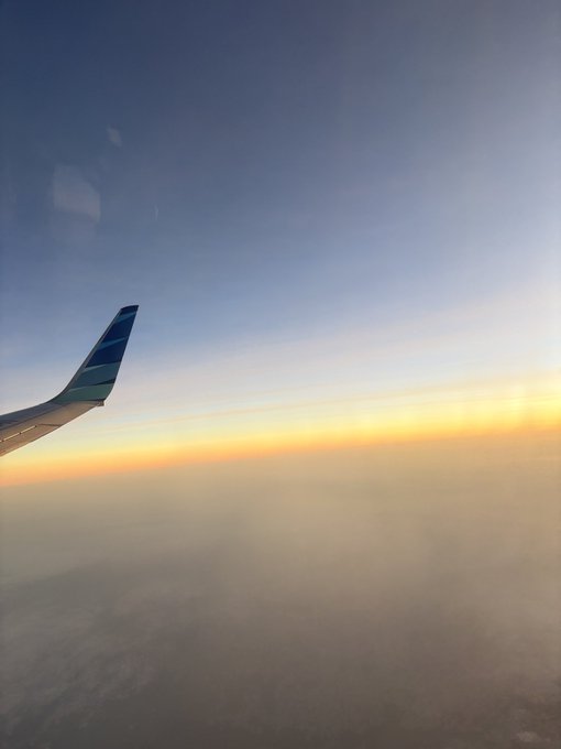 A view from an airplane window at sunset, showing the wing of a Garuda Indonesia aircraft (recognizable by its blue and teal tail livery) soaring above a blanket of clouds with a golden horizon glowing in the distance. The serene sunset scene captures the peaceful end of a journey home from Bali after attending CoinfestAsia, reflecting the transition from conference travel back to focused work.