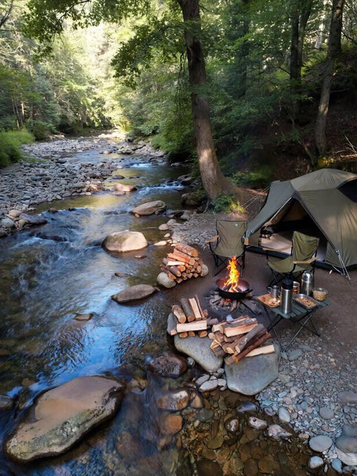 A serene campsite by a rocky stream with a green tent, a campfire surrounded by stacked firewood, and two camping chairs in a lush forest.