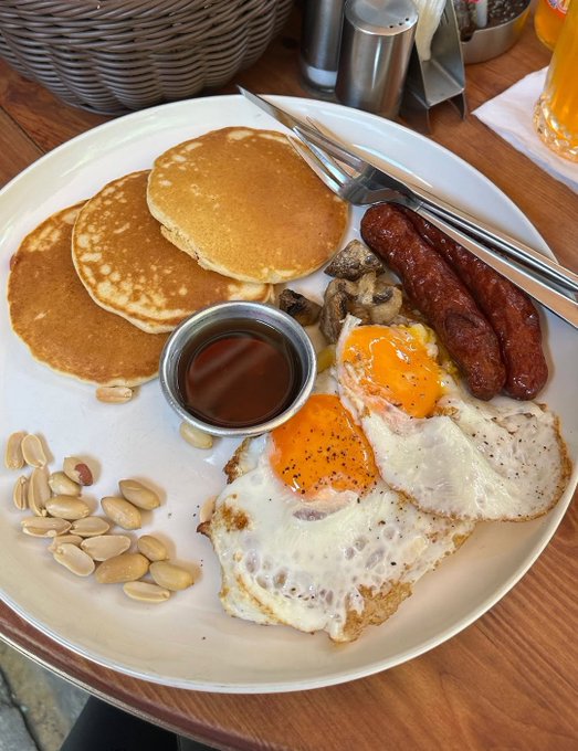 A hearty breakfast plate featuring pancakes, fried eggs, sausage links, sautéed mushrooms, peanuts, and a small cup of syrup, served on a wooden table. The image's leisurely weekend brunch vibe contrasts with the post's message about crypto security and self-custody via @Ledger, playfully tying the "not your keys, not your coins" mantra to the visual reminder that breakfast is best enjoyed without worrying about losing your stack.