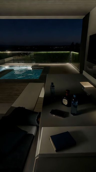 A dimly lit outdoor patio or balcony area at night with a hot tub, lounge seating, and a few water bottles resting on a table.