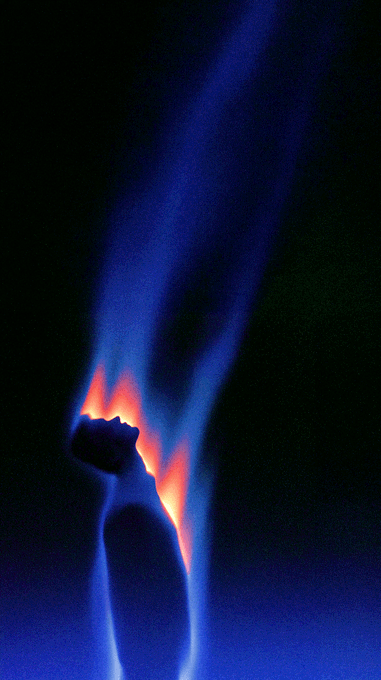 The image features a blue flame coming out of an object, possibly a candle or a lighter. The flame has a bright orange hue that stands out against the dark background. The photo appears to be taken at night with the flame being the main source of light in the scene. There is also text present in the image which reads "Gm Auction is live! 7 hours left, 0.015 ETH". This suggests that there might be an auction happening or a sale taking place related to candles or similar items.