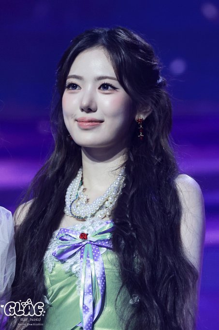 A young woman with long dark hair smiles slightly, wearing a green and purple dress with a large pearl necklace and cherry earrings, with a 'GLAD' watermark visible.