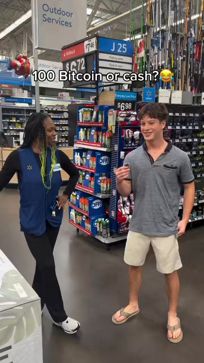 The main focus of the image is a man standing next to a woman. They both seem to be engaged in conversation while shopping at a store. In front of them, there's an item that they're looking at together, possibly discussing its features or price. A computer monitor can also be seen in the background on one side of the image. The post appears to be directed towards the woman and talks about Bitcoin as payment options for the item she is holding. It seems like the man might have made a comment about Bitcoin being 100 Bitcoins or $20,000 cash for that item.