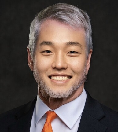 A professional headshot of a smiling East Asian man with graying hair and a short beard, wearing a dark suit, light blue shirt, and an orange tie against a black background. The polished corporate portrait style conveys professionalism and confidence, complementing the post's message about steady, confident deployment of capital.