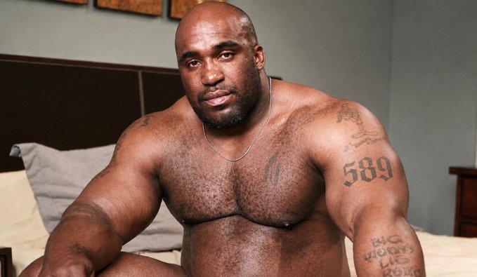 A muscular, bald man with a beard sits shirtless on a bed, displaying visible tattoos including "589" on his upper arm and text reading "BIG WOLF LIKES ROAD" on his forearm. The image humorously accompanies the caption about XRP's price, with the man's muscular build and the "589" tattoo visually playing on the "$2" price reference.