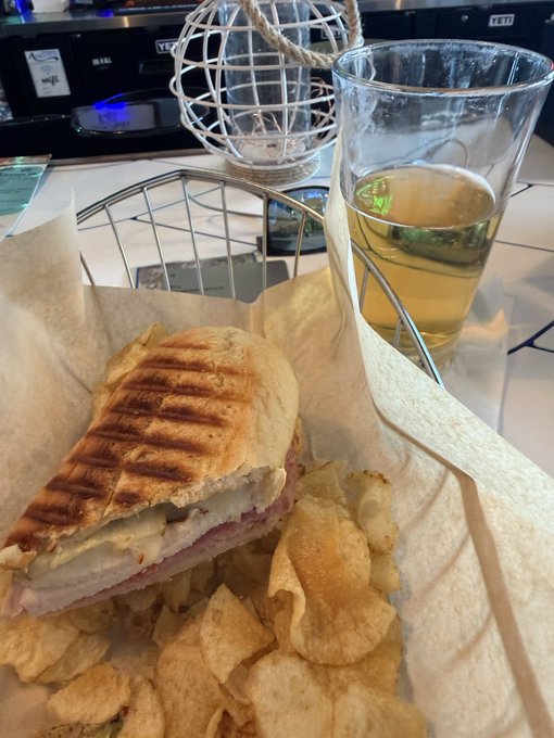 A pressed Cuban sandwich with visible grill marks sits on parchment paper next to a pile of pork rinds and a plastic cup of light-colored beer.