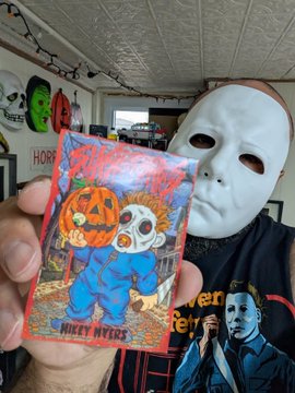 A hand holds up a colorful trick-or-treat candy bag featuring a pumpkin-headed character, displayed against a background with a white mask and horror-themed decor.