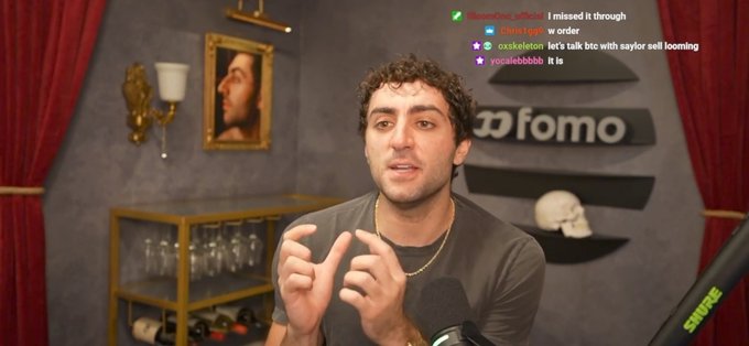 The image shows a curly-haired man in a gray shirt speaking into a Shure microphone in a dimly lit studio decorated with red curtains, a portrait painting, wine bottles, and a "fomo" sign, while Twitch chat messages reference "saylor" in the corner. The setup and chat interaction relate to the post's description of a streamer/content creator engaged in a live conversation with his audience.