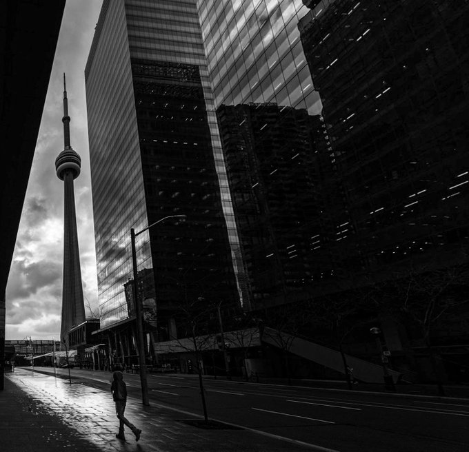The image shows a person walking down an empty city street, which has tall buildings with reflective windows surrounding them. The scene takes place during the evening or nighttime, as indicated by the lack of activity and the darkness. In the background, there is a towering structure that stands out against the night sky. This building could be part of a skyscraper in a downtown area.