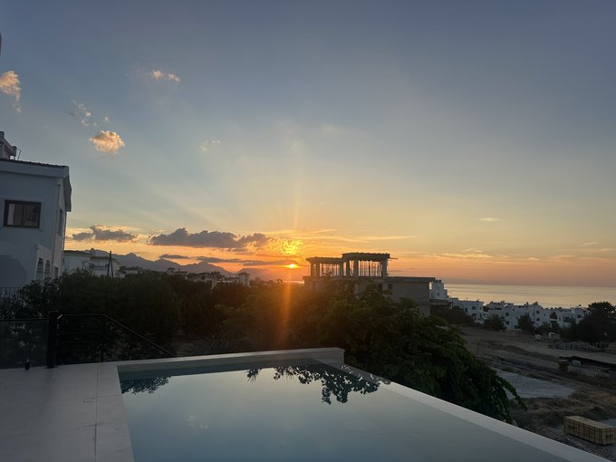 A scenic sunset view over a coastal area with an infinity pool in the foreground, featuring white buildings and ancient ruins silhouetted against the golden sky.