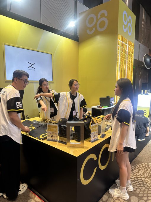 A trade show or convention booth with yellow branding displaying the number '96', where several people in matching branded apparel are interacting at a counter showcasing products.
