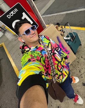 A selfie of a person wearing a colorful graffiti-patterned jacket and sunglasses, standing near a door sign and luggage in what appears to be a parking garage.