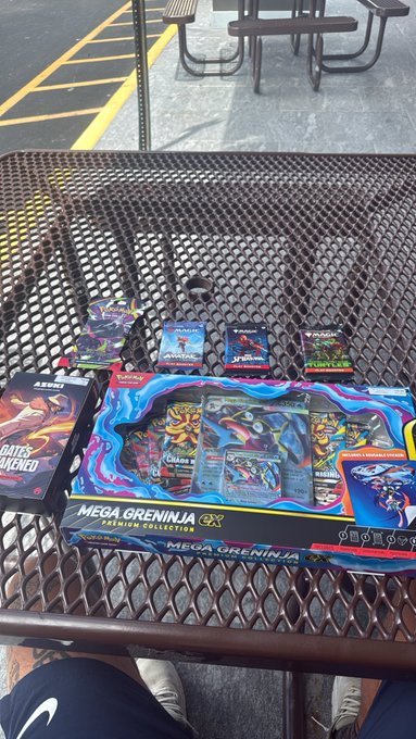 Pokémon trading card products including a 'MEGA GRENINJA GX Premium Collection' box and several booster packs (including 'Astonish' and 'Hunter') arranged on a metal mesh table outdoors.