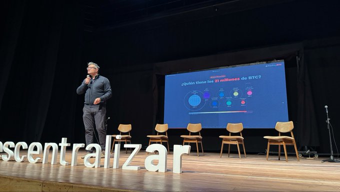 A man speaks on stage at what appears to be a 'descentralizar' conference, standing next to a presentation slide asking '¿Quién tiene los 21 millones de BTC?' (Who has the 21 million BTC?) with chairs lined up beside him.