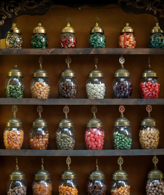 Shelves filled with decorative glass jars with ornate brass lids containing various colored candies, sweets, and treats in greens, reds, oranges, pinks, browns, and creams.