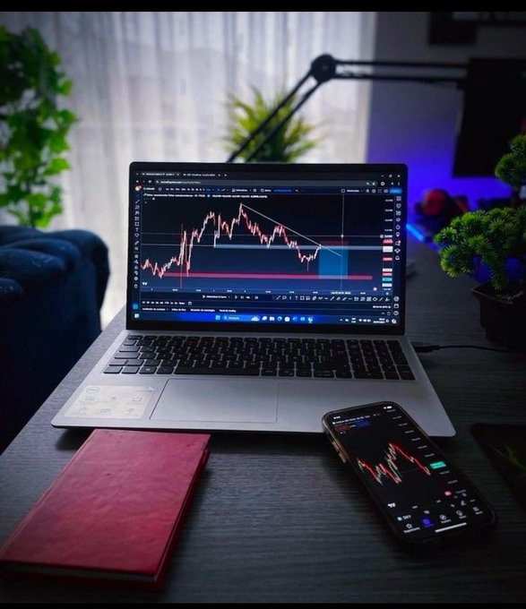 A desk setup featuring a laptop displaying a trading chart with red candlestick patterns, a smartphone also showing a trading chart, a red notebook, and ambient lighting with plants in the background.