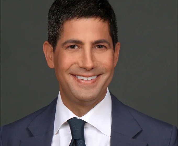 A professional headshot of a smiling man in a dark suit and tie against a gray background.