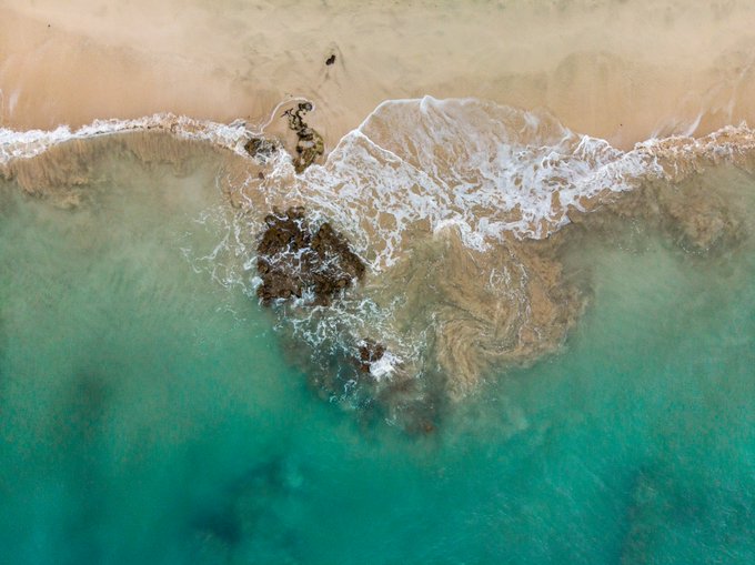An aerial view captures turquoise ocean waves crashing against a sandy beach, with rocks and white foam visible at the shoreline.