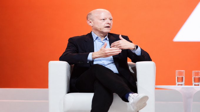 A bald man in a dark blazer and light blue shirt sits in a white chair against an orange backdrop, gesturing with his hands as he speaks during what appears to be a panel discussion.