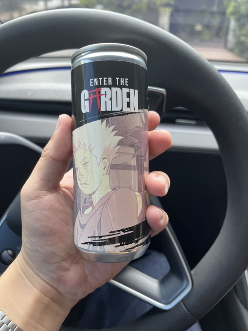 A hand holds a tall black energy drink can branded "ENTER THE GARDEN" featuring anime-style artwork, photographed inside a Tesla vehicle with the steering wheel visible in the background.