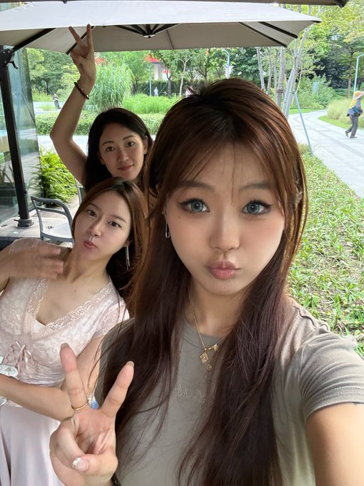 Three young women take a selfie outdoors under a canopy, with the front woman making a peace sign and a playful expression while the others pose behind her.