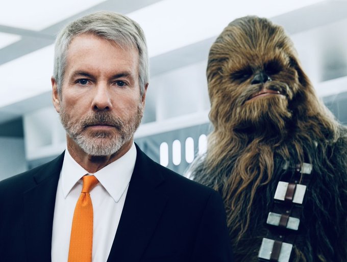 The image features a man wearing a suit and tie standing next to a large Chewbacca statue with long brown hair, which appears to be posed for a photo. The man's face can be seen clearly as he looks directly at the camera. The background of the image is blurred, drawing attention to both the man and the Chewbacca statue.