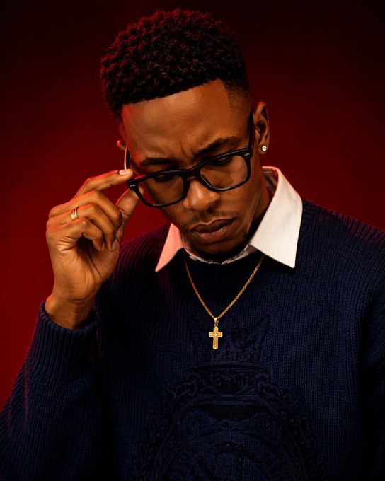 A man with short curly hair adjusts his glasses while wearing a navy blue sweater, a white collared shirt, and a gold cross necklace against a red background.
