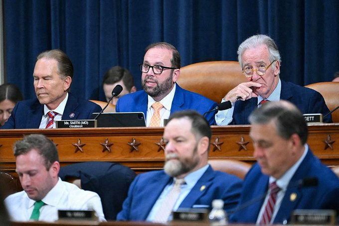 A congressional hearing with several men in suits seated at a panel, including one man with glasses speaking at a microphone, with nameplates visible on the desk.