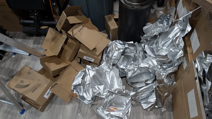 A cluttered storage area filled with discarded cardboard boxes, silver padded mailers, and an air compressor.