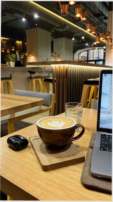 A coffee shop scene with a latte featuring a leaf pattern in a brown cup on a wooden coaster, sitting next to a laptop, earbuds case, and glass of water.