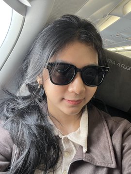 A selfie of a woman with long dark hair wearing black sunglasses and a brown jacket, seated inside an airplane with the headrest text 'RIA ATLUI' partially visible.